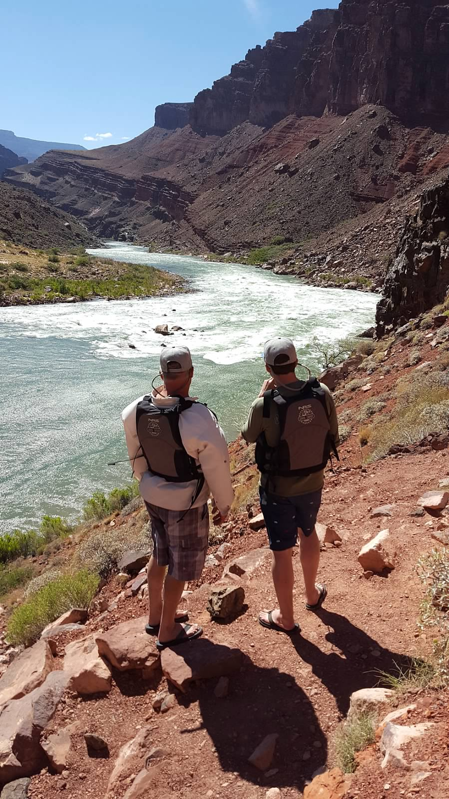 Two men are standing on a trail looking at a river.