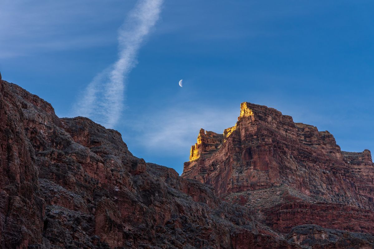 Red rock canyon with blue sky, a crescent moon, and a contrail.