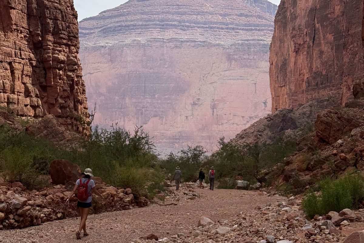 Hikers in a canyon, walking on a rocky path. Red rock walls surround the path.