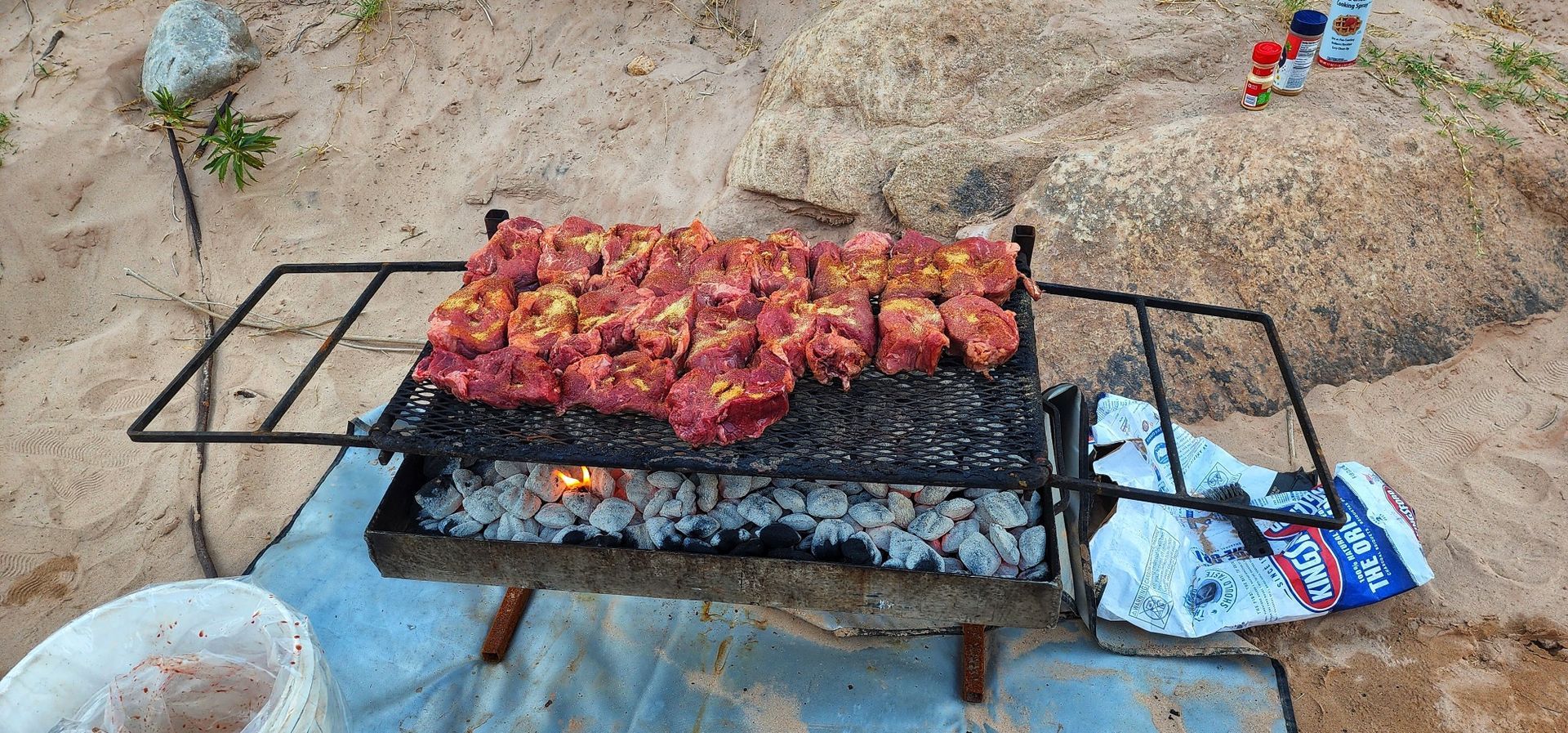 A grill filled with meat is sitting on top of a blue blanket on the beach.