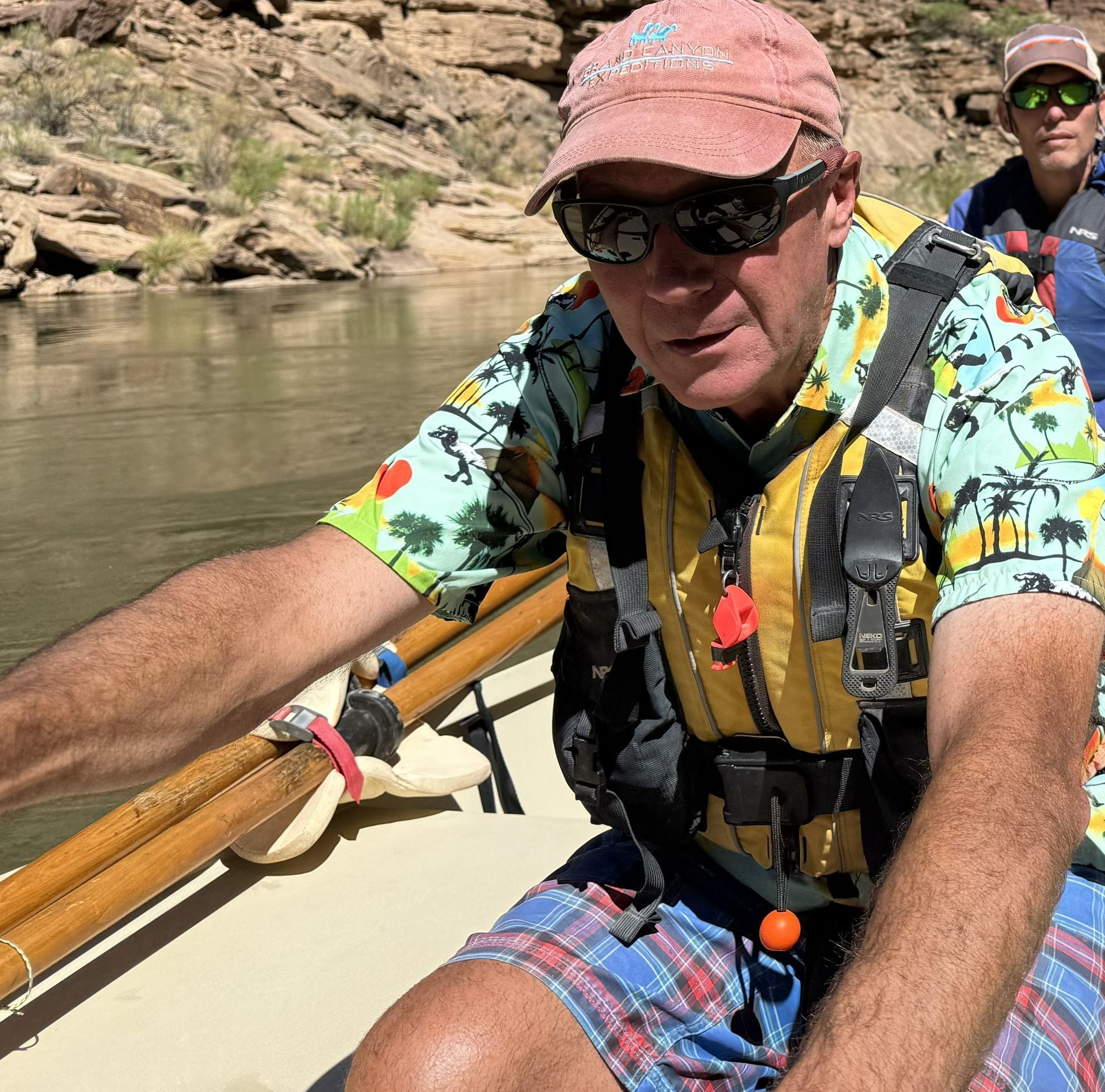 Man in sunglasses and hat rows a raft on a river, wearing a life vest and colorful shirt.