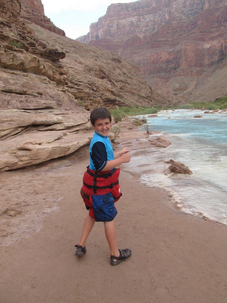 A young boy wearing a life jacket is standing on a beach near a river.