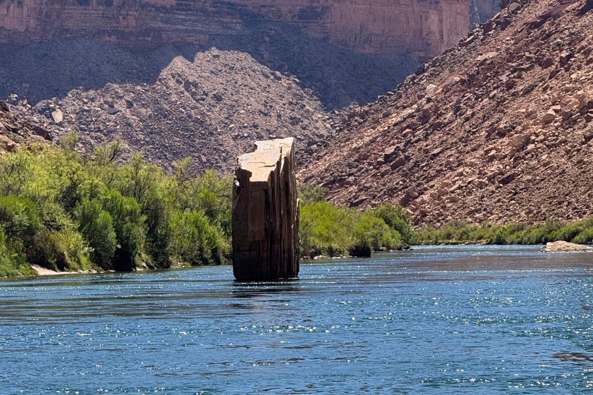 A tall, weathered rock formation stands in a blue river. Lush green trees line the banks. Mountains in the background.