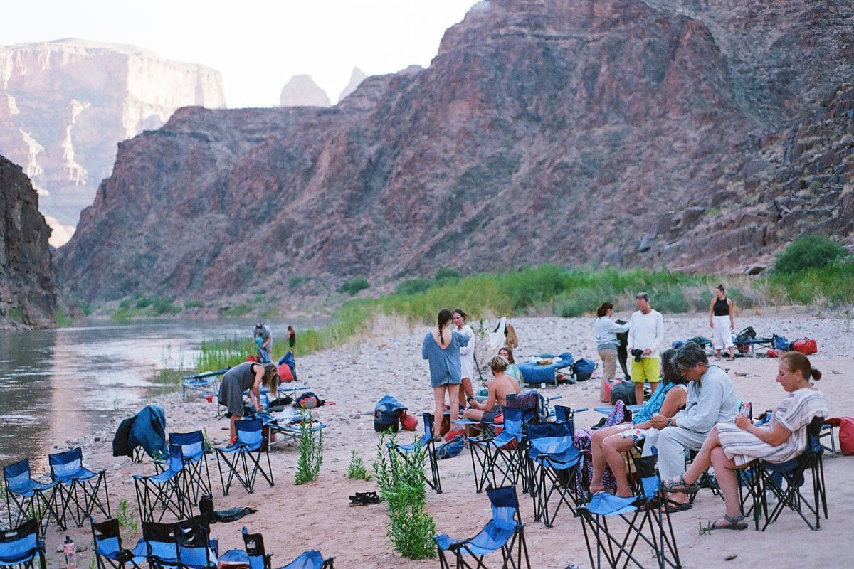 People gathered at a riverside campsite with folding chairs, mountains in background, early evening light.