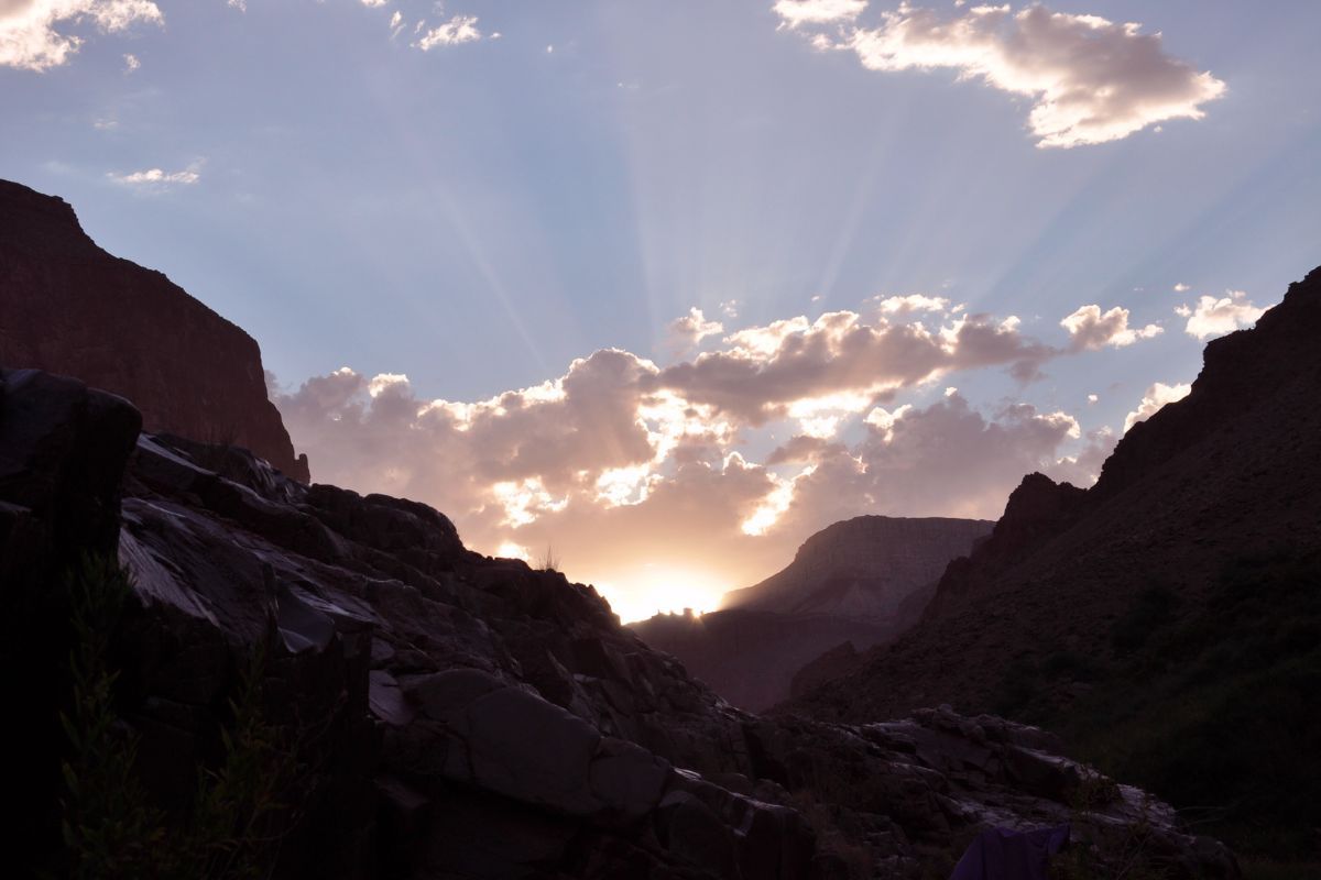 Sun setting over a canyon, illuminating clouds with golden light; rocky landscape in silhouette.