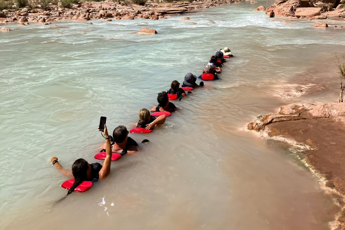 People floating in a line on red kickboards down a milky river, near a reddish-brown bank.