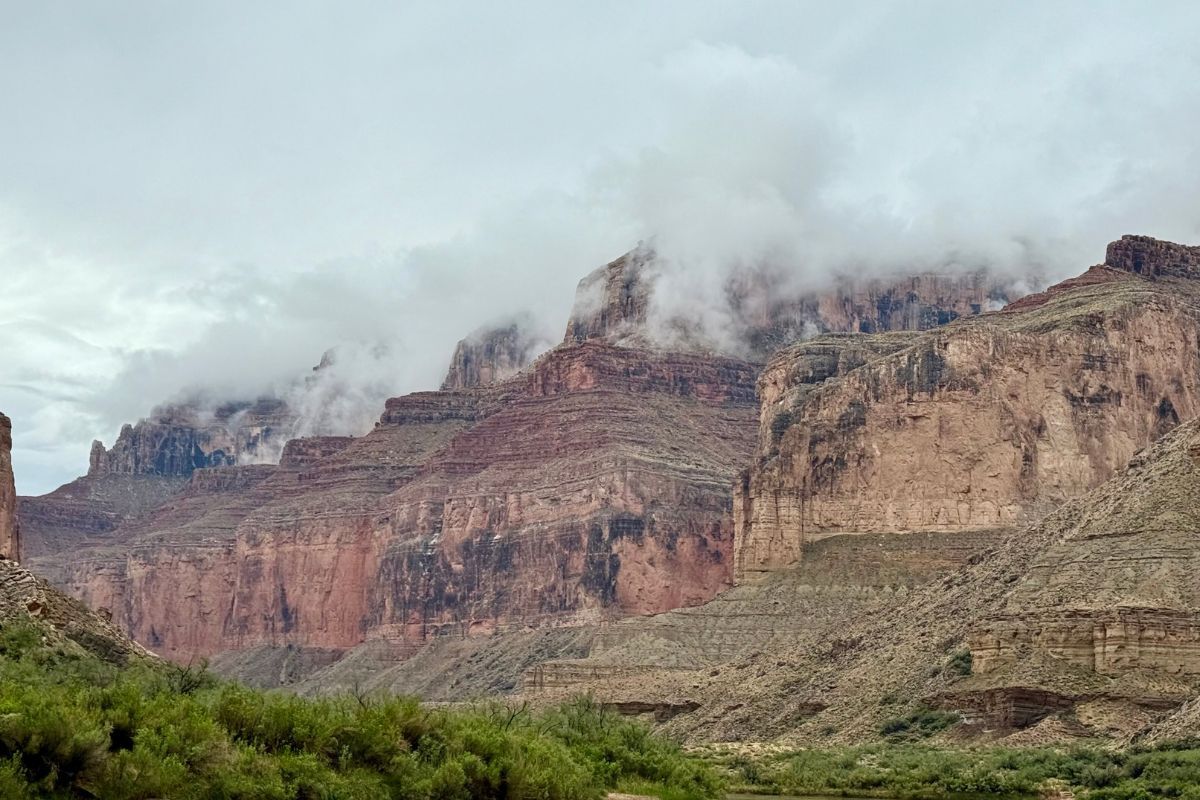 Red and tan canyon walls with low-hanging clouds. Green vegetation in the foreground.