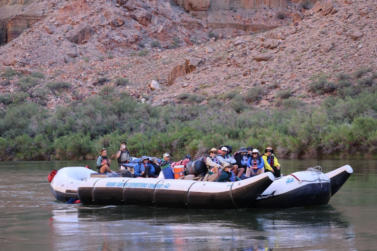 Raft with passengers on a river, cliffs in the background. People seated, gear visible.