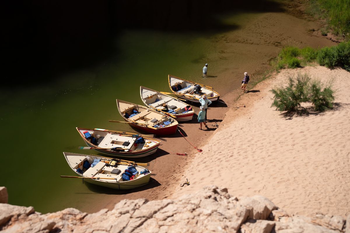 Boats lined up on a sandy riverbank, with people near the water and a view of the canyon.