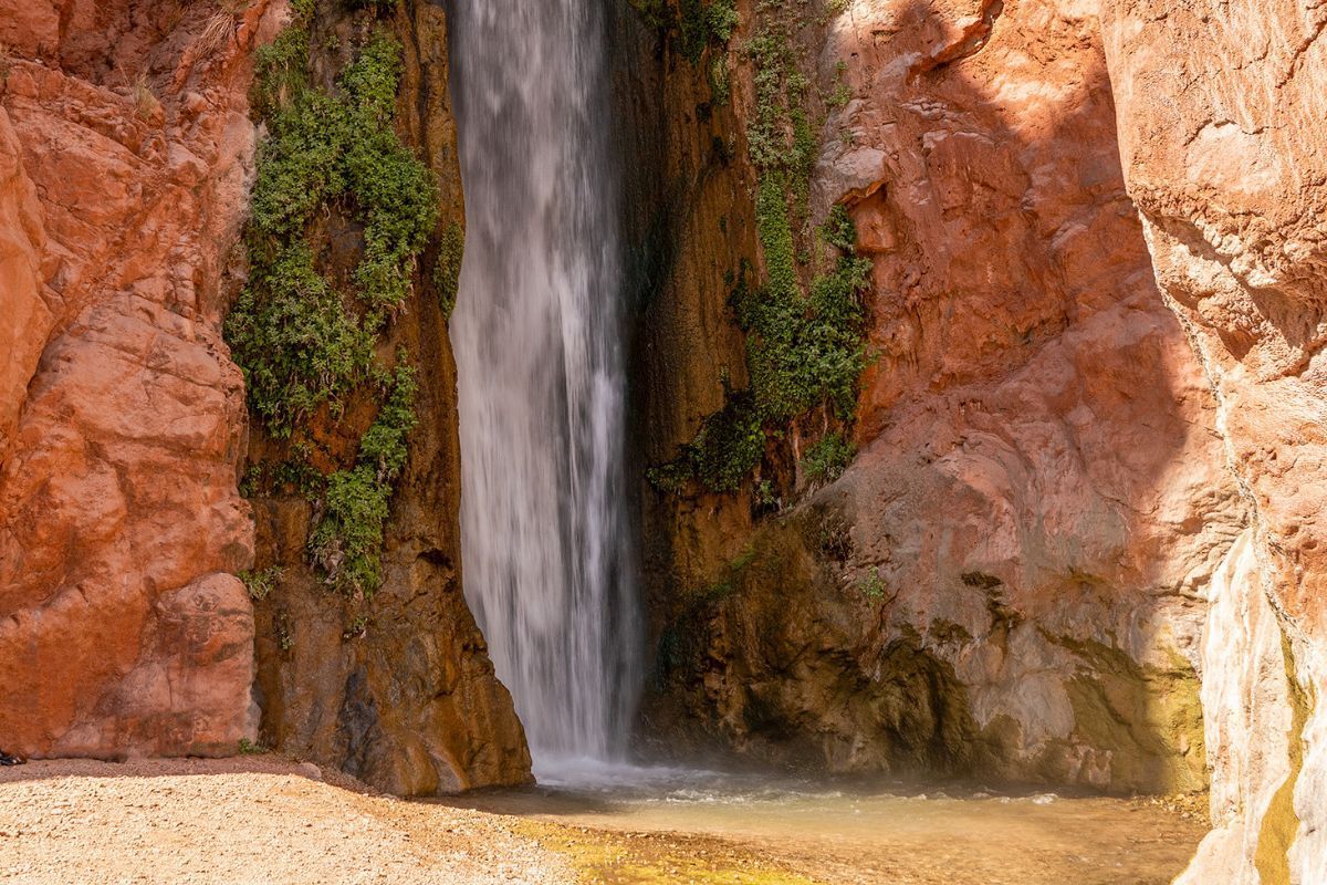 Waterfall cascading down red rock canyon, small pool at base.