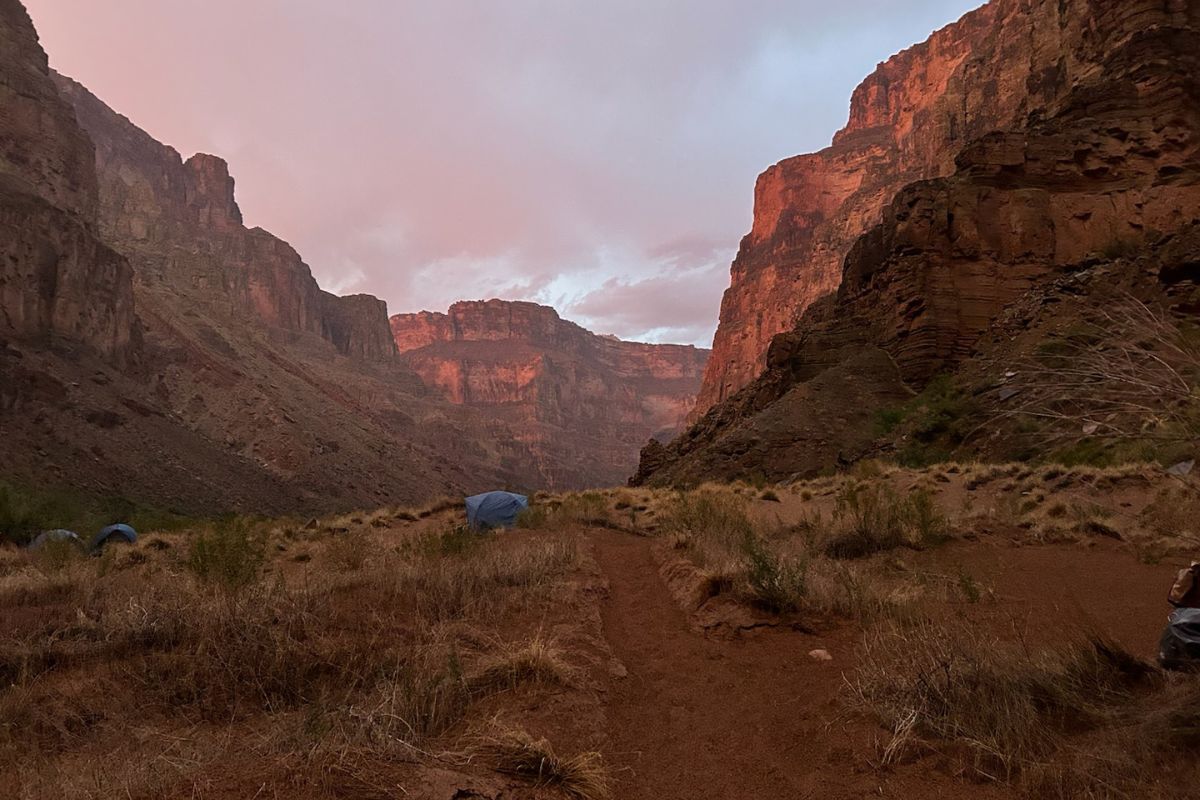 Canyon view at sunrise with tents. Pink sky, red rock walls, path, and brown vegetation.