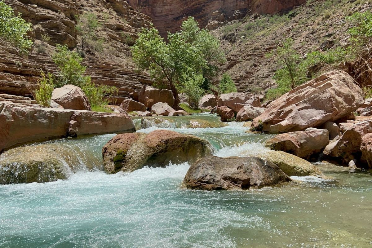 Fast-flowing turquoise water cascades over rocks in a canyon, surrounded by red-brown rock walls and sparse greenery.