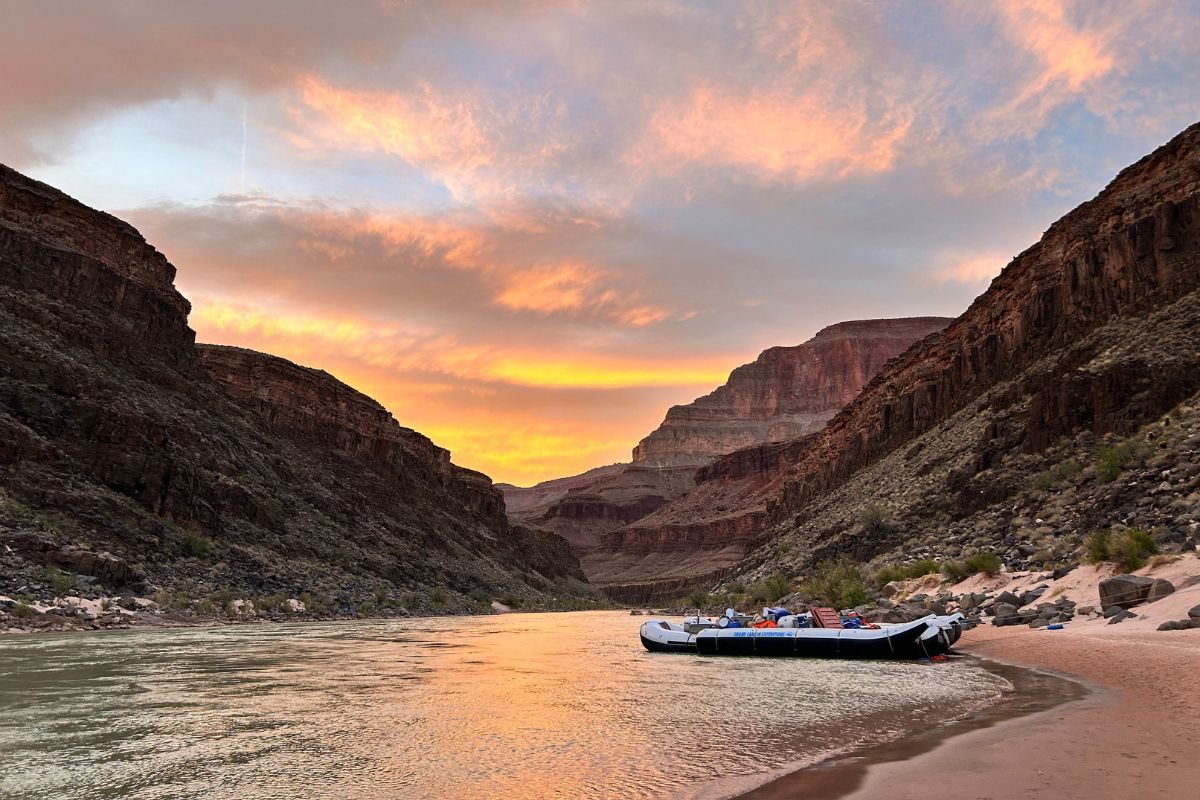 Raft on shore in a canyon at sunset; golden sky, brown cliffs, calm water.