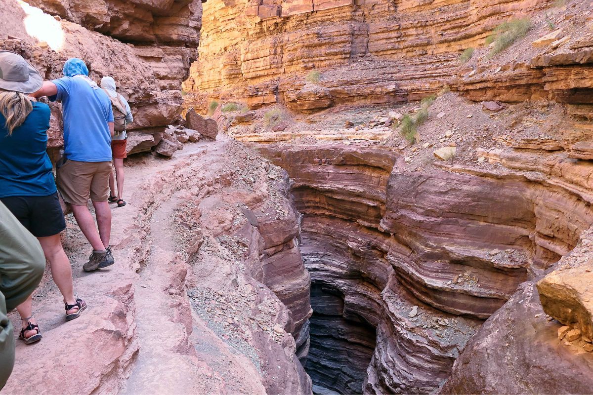 Hikers in a narrow desert canyon with layered rock formations and a deep, dark opening.