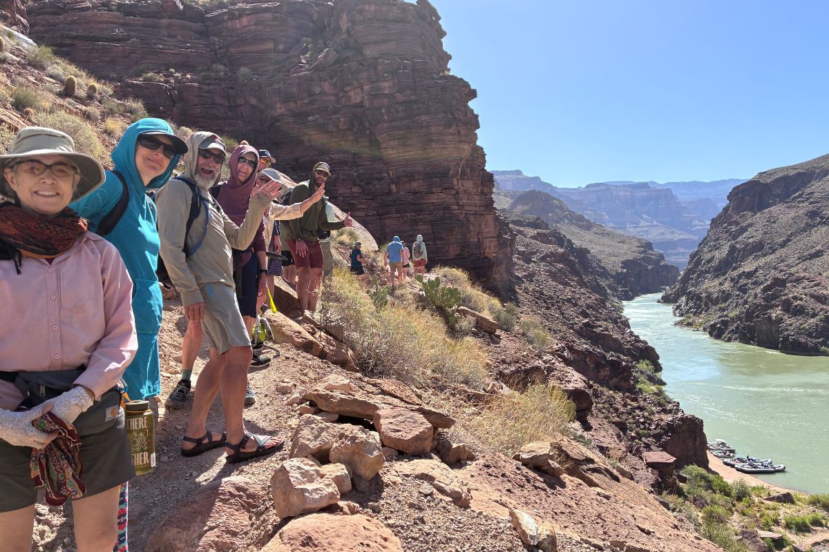 Group hikes along a cliffside trail overlooking a river in a canyon. Sunny day, blue sky.