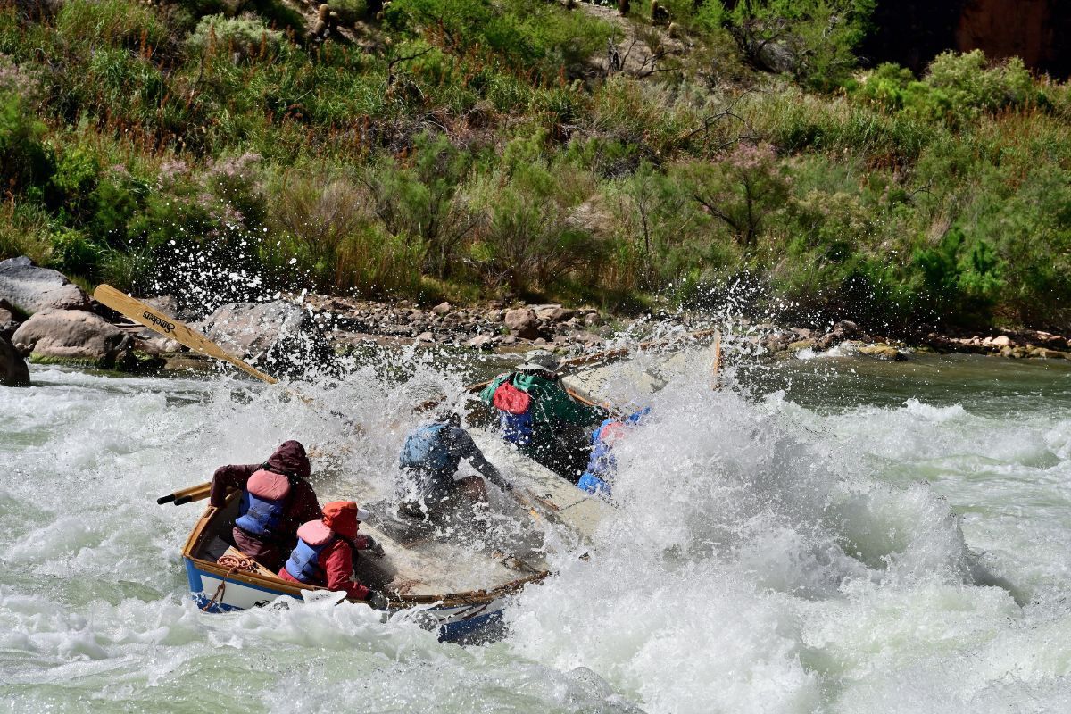 Rafting boat navigating turbulent rapids, white water splashing. Green foliage in background.