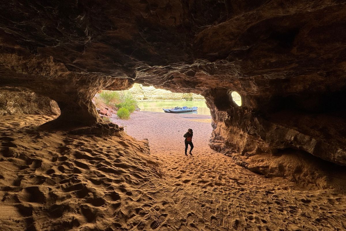 Person stands in cave entrance, looking at boat on a dry riverbed.