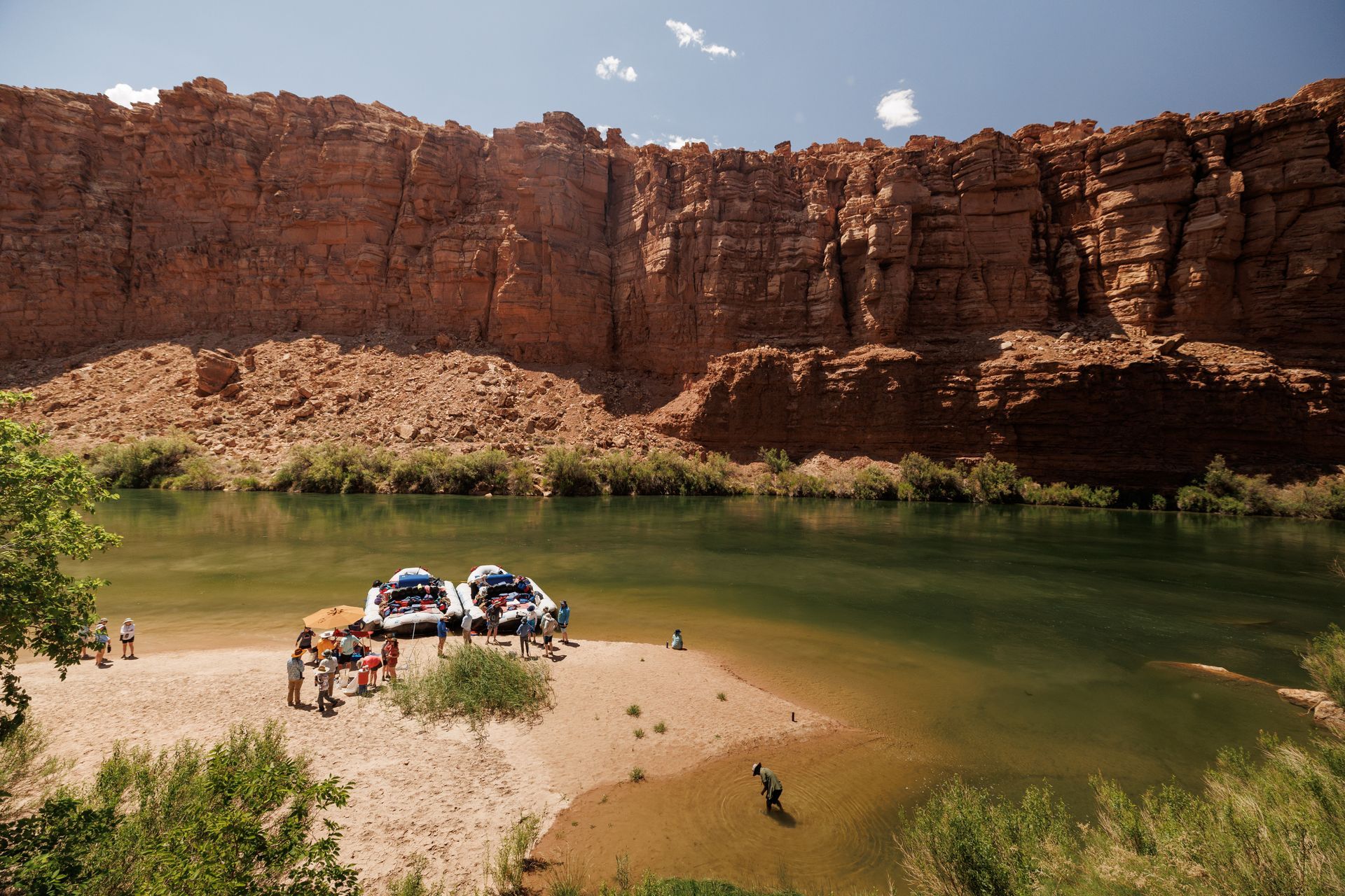 People near rafts on a riverbank, red rock canyon backdrop, sunny day.