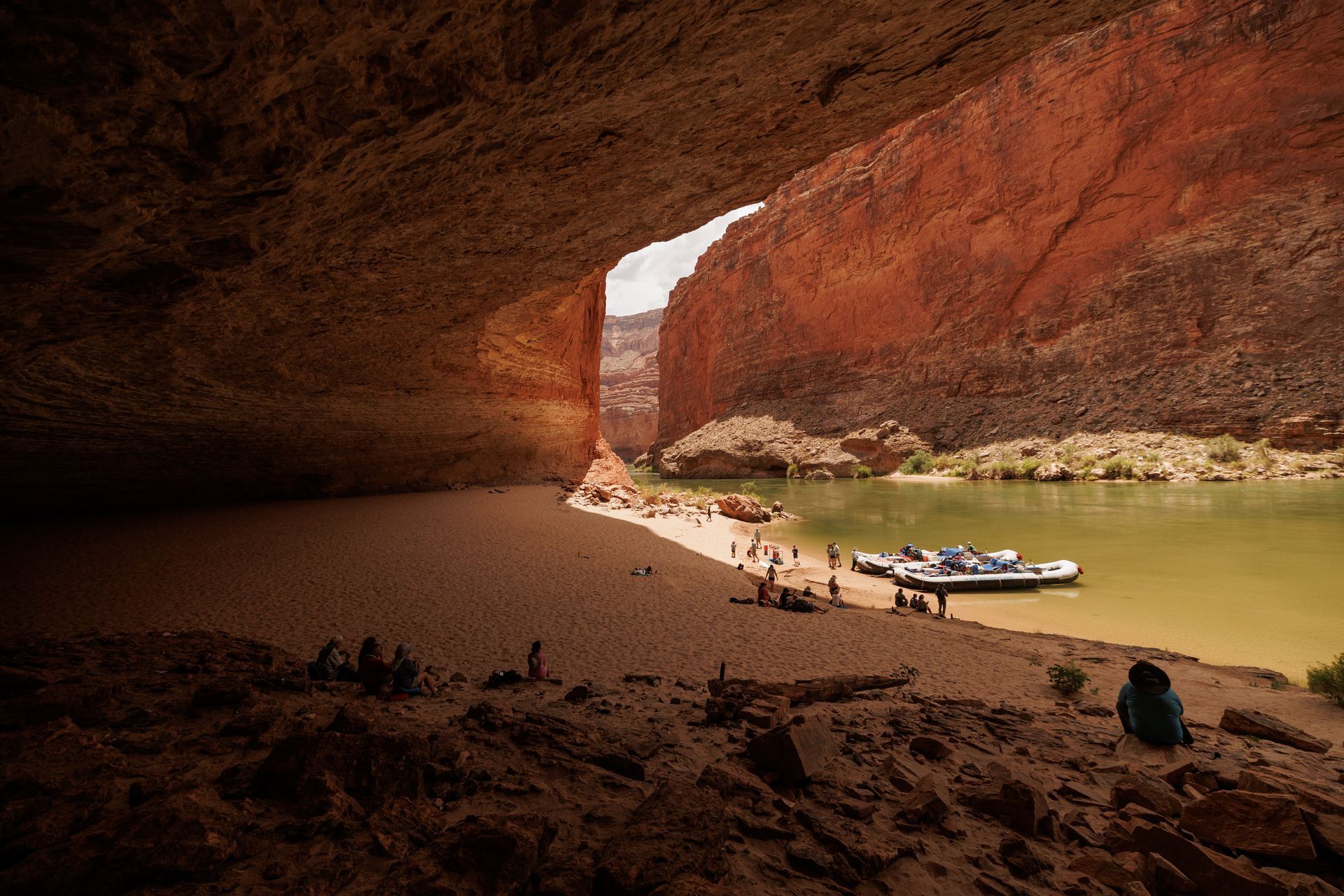 Cave opening onto a sandy beach, river, and rafts. Red rock walls frame the scene. People are present.