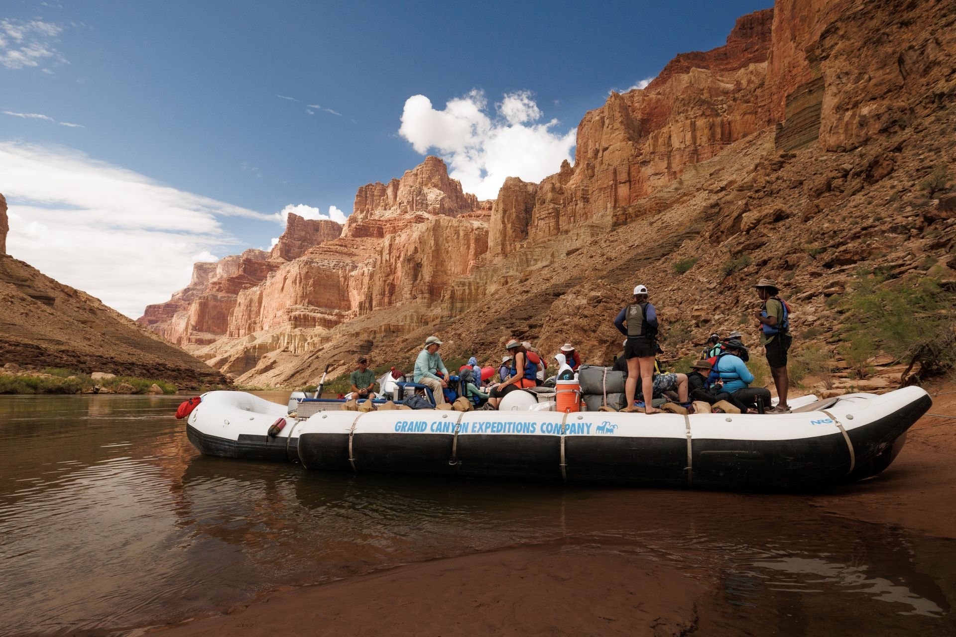 A raft with people on a river next to a red rock canyon under a blue sky.