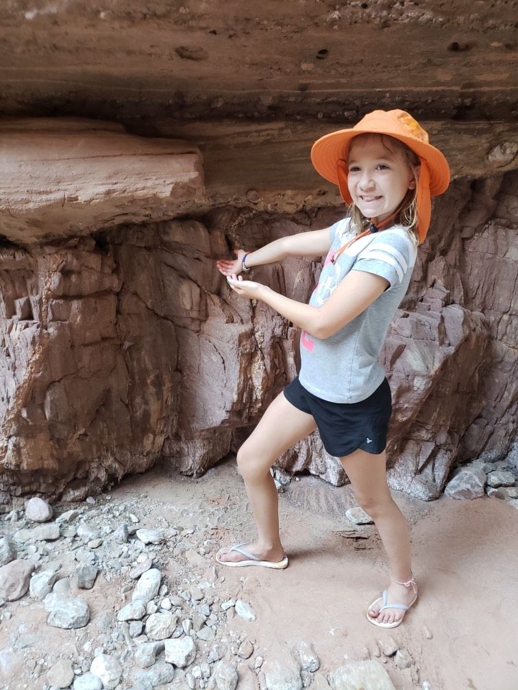 A little girl wearing an orange hat is standing in front of a rock wall.