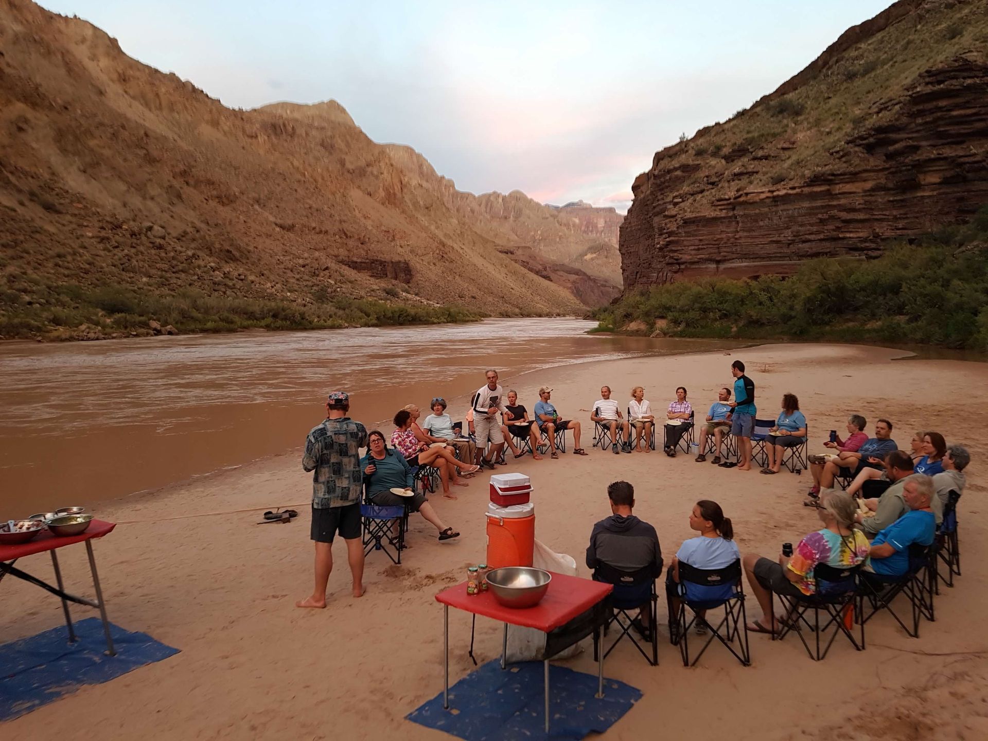 A group of people are sitting in a circle on the beach near a river.