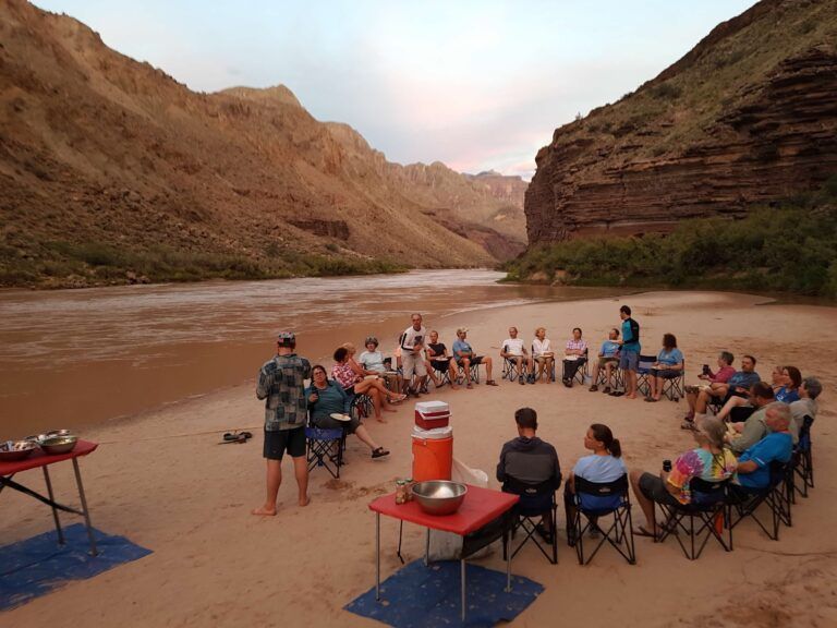 A group of people are sitting in a circle on the beach near a river.