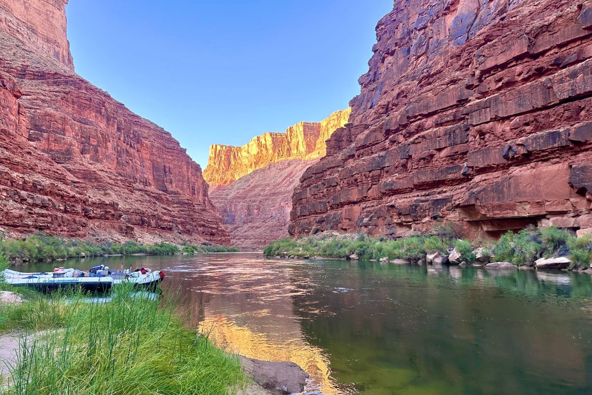 River flowing between red canyon walls; boats docked near green vegetation.