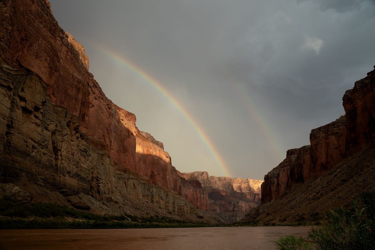 A rainbow is visible over a river in a canyon.