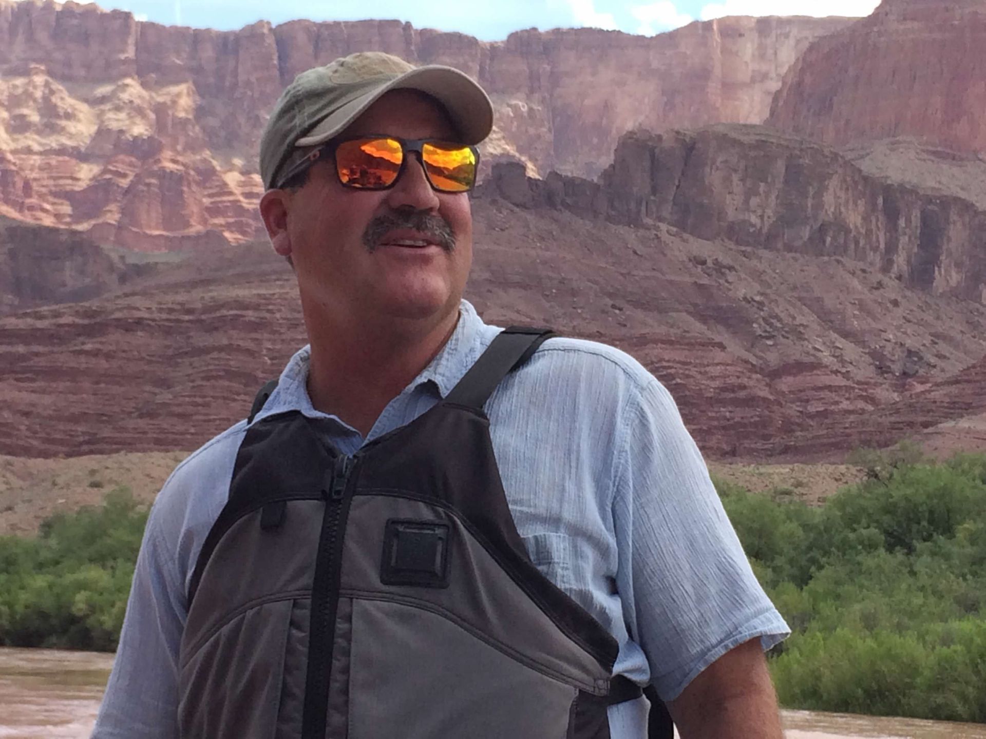 A man wearing sunglasses and a hat is standing in front of a mountain.