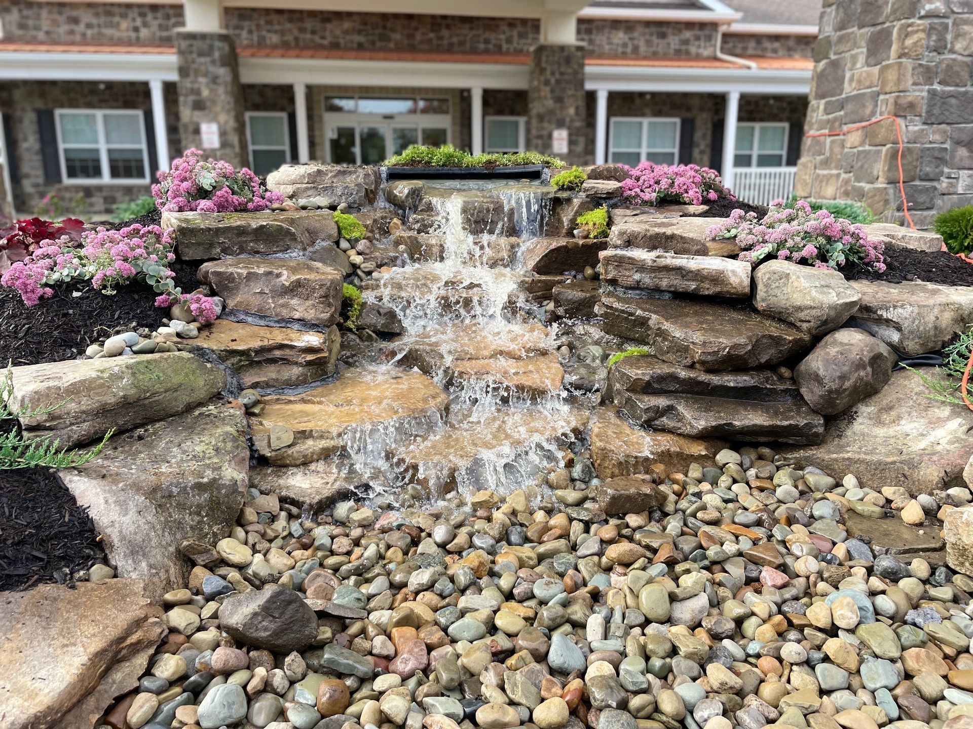 A waterfall is surrounded by rocks and flowers in front of a house.