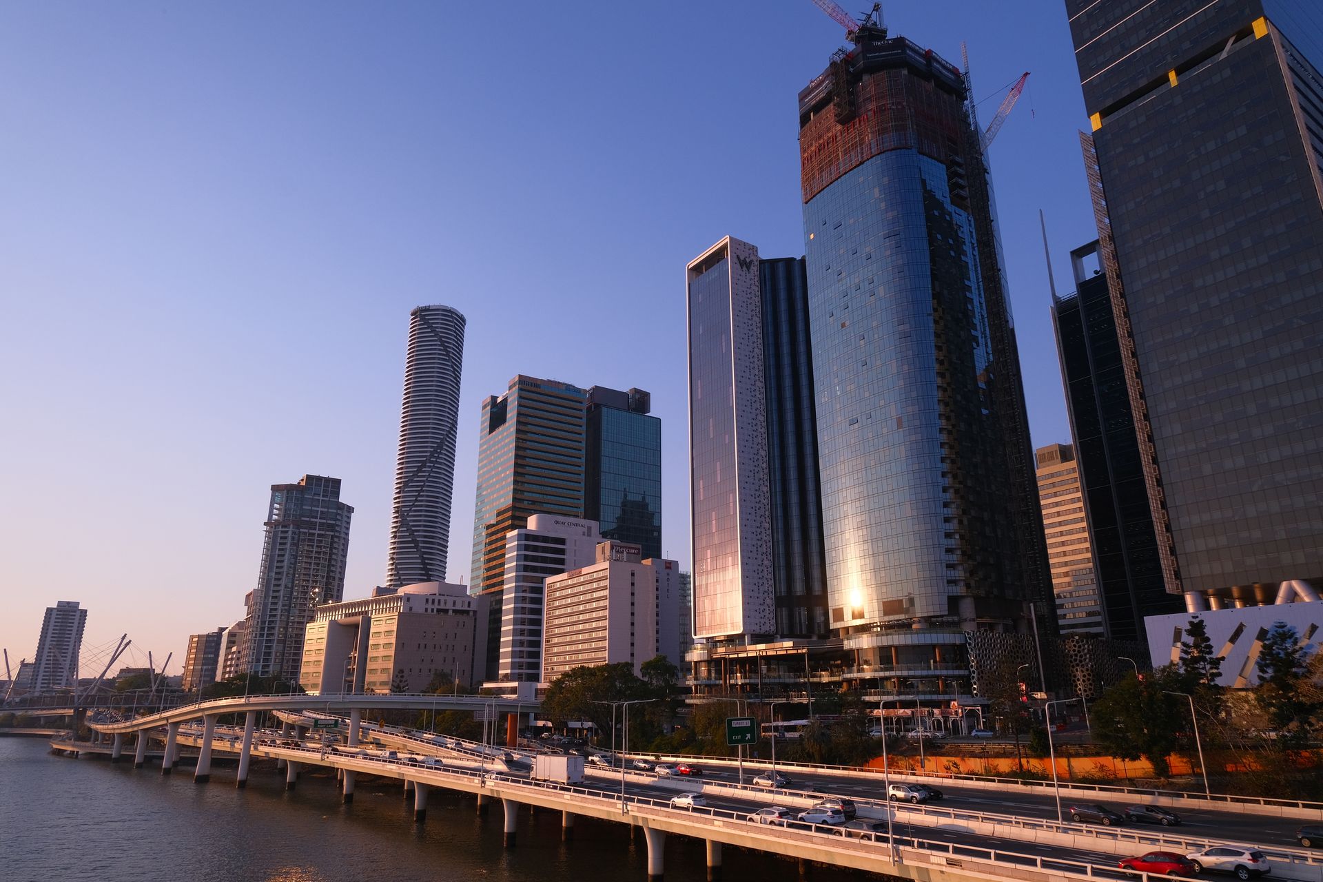 City skyline with tall buildings under construction, reflecting sunlight. A bridge with traffic is in the foreground, and a clear sky is overhead.