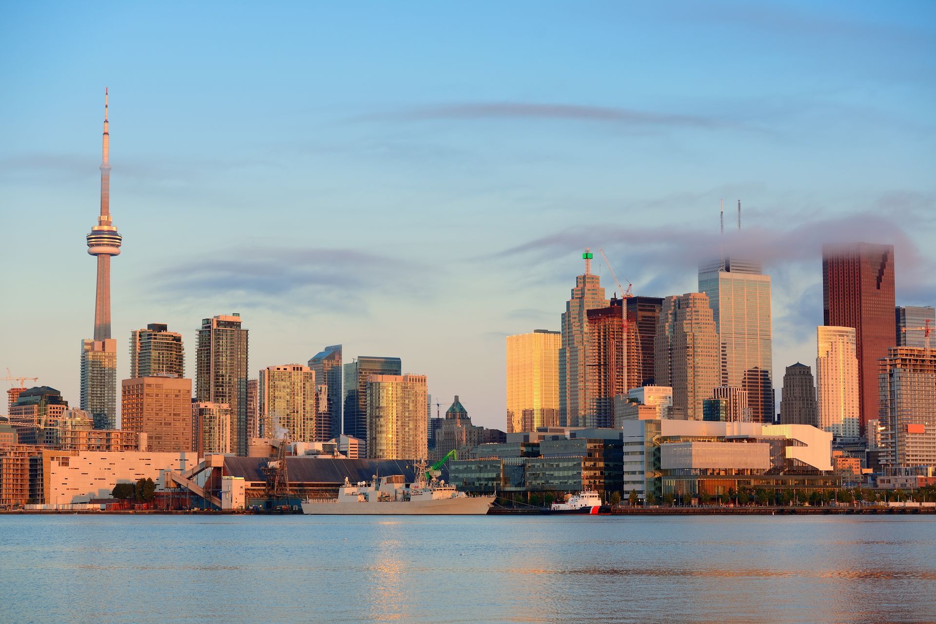 Toronto skyline at sunrise, featuring the CN Tower and other skyscrapers reflecting the warm light over the lake.