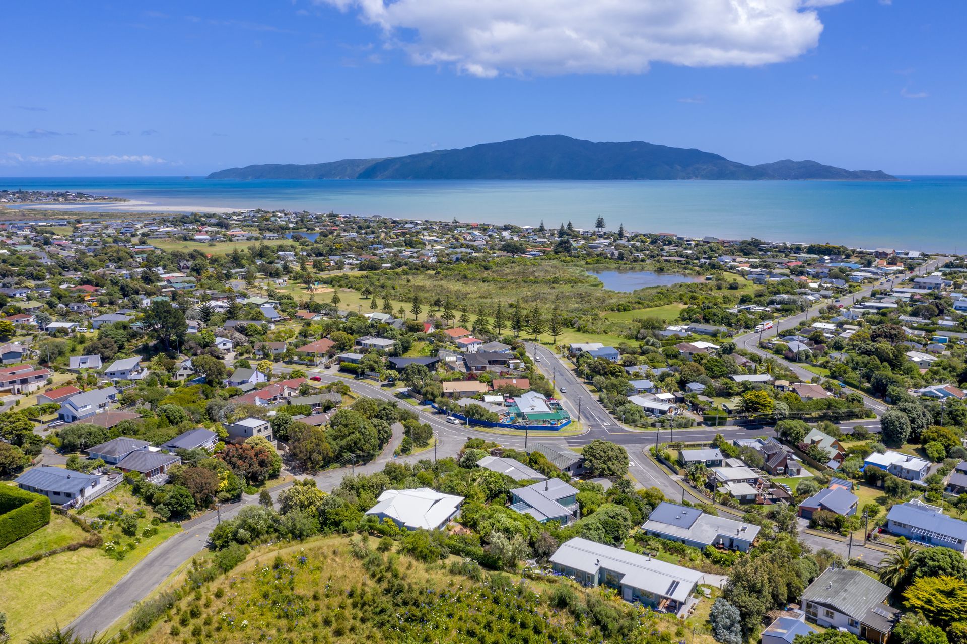Aerial view of a coastal town with houses, a lake, and a large mountain in the background under a blue sky.