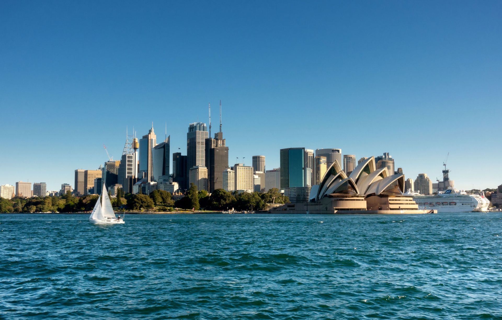 Sydney skyline with the Opera House on a sunny day, sailboat in foreground, and blue water.