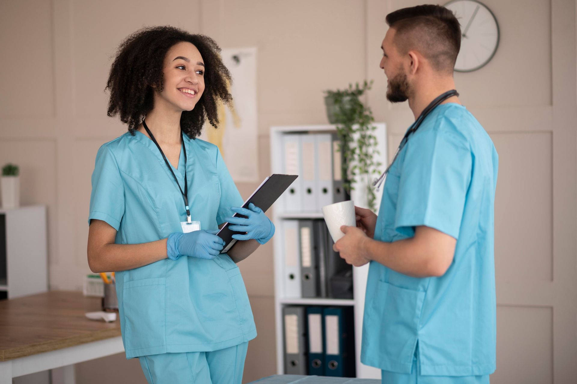 Two healthcare workers in blue scrubs smile while conversing in an office. One holds a tablet and wears gloves.