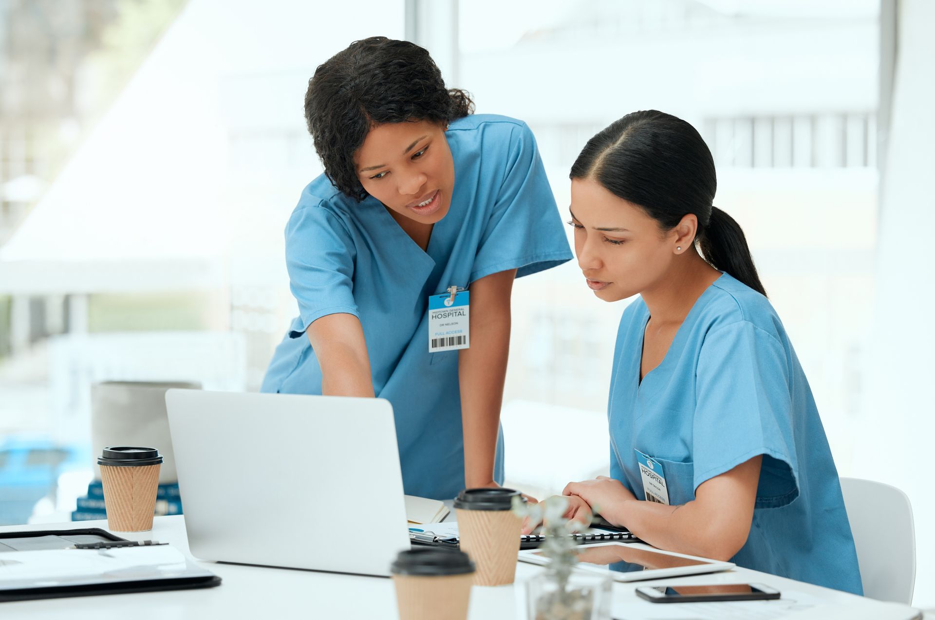 Two nurses in blue scrubs, looking at a laptop in an office setting. One points at the screen, the other looks intently.