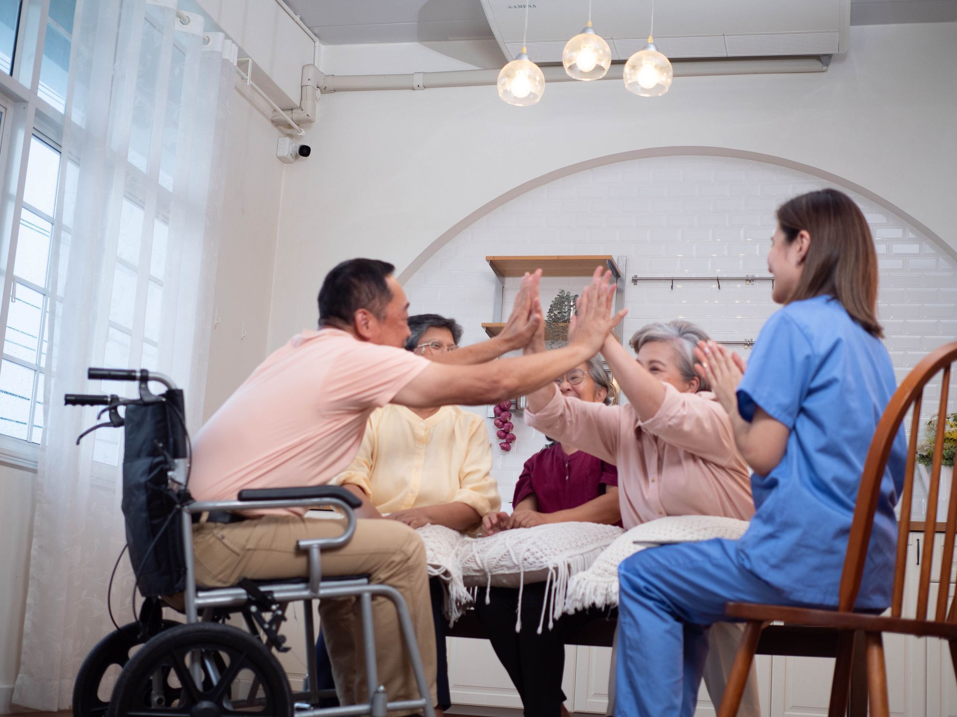 A group of seniors and a nurse in a bright room, high-fiving. A man sits in a wheelchair.