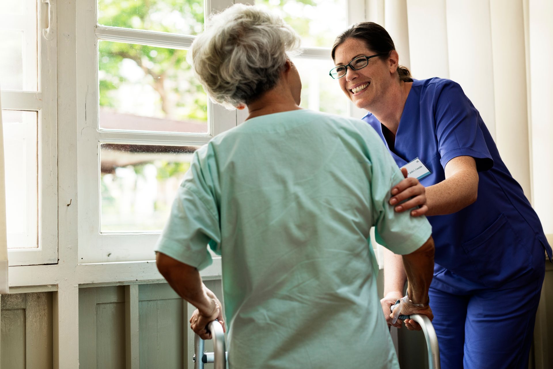 A healthcare worker in blue scrubs assists an elderly patient in a hospital gown using a walker near a window. Both are smiling.