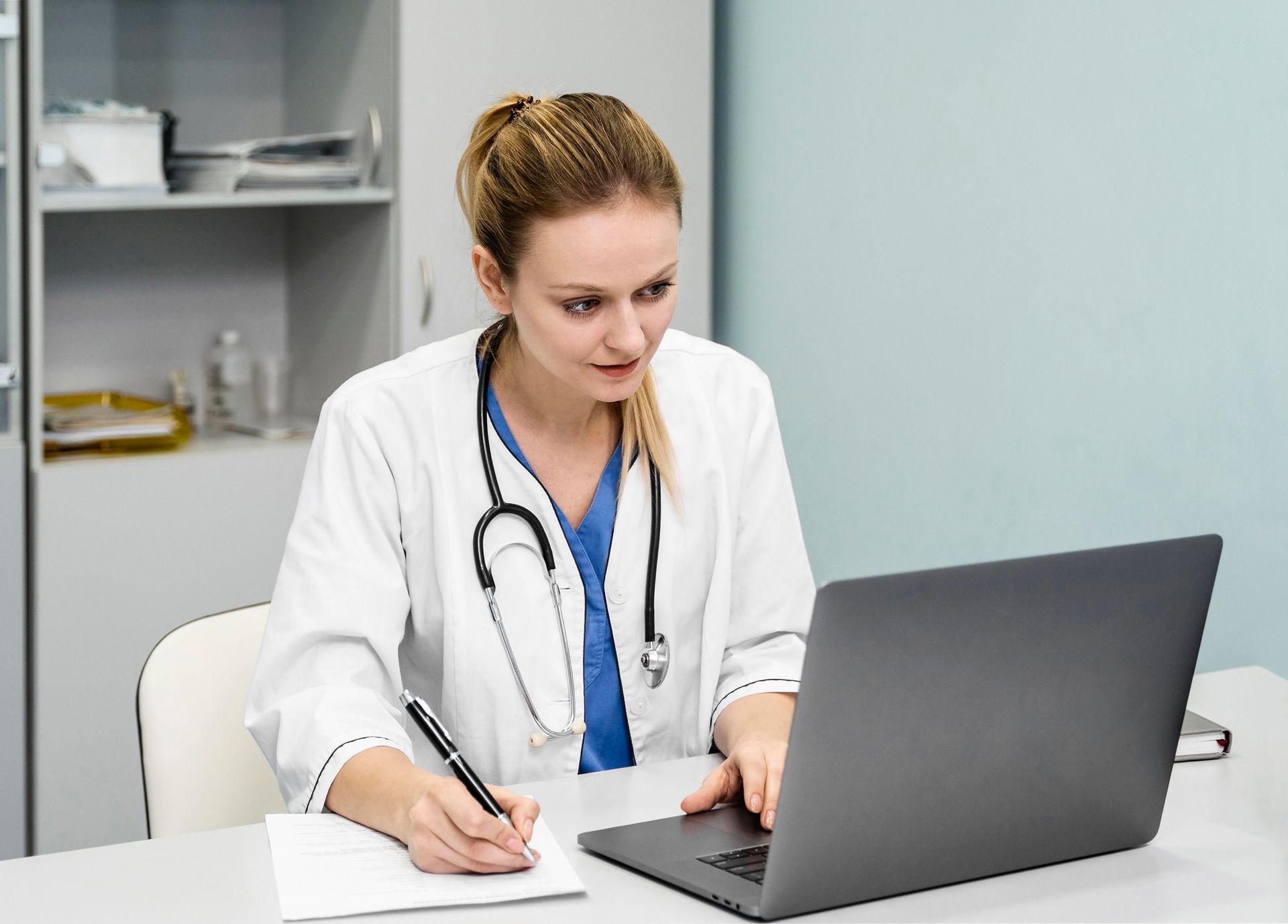 A female doctor in a white coat with a stethoscope, looks at a laptop screen while taking notes in a medical office.
