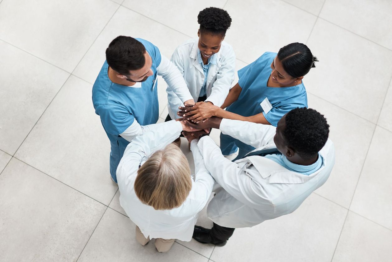 Medical team in scrubs and lab coats, hands stacked together in a circle, celebrating in a brightly lit room.