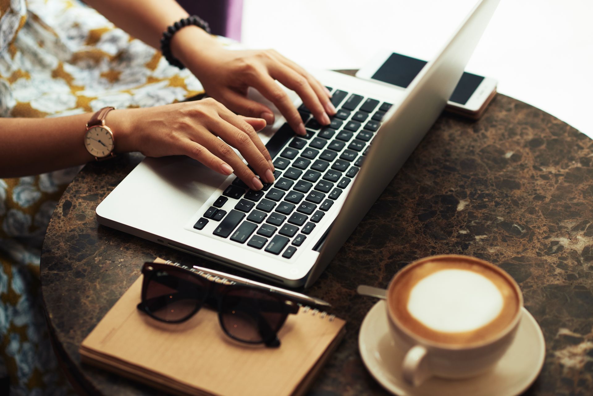 Person typing on laptop at a cafe table with coffee, notebook, and sunglasses.