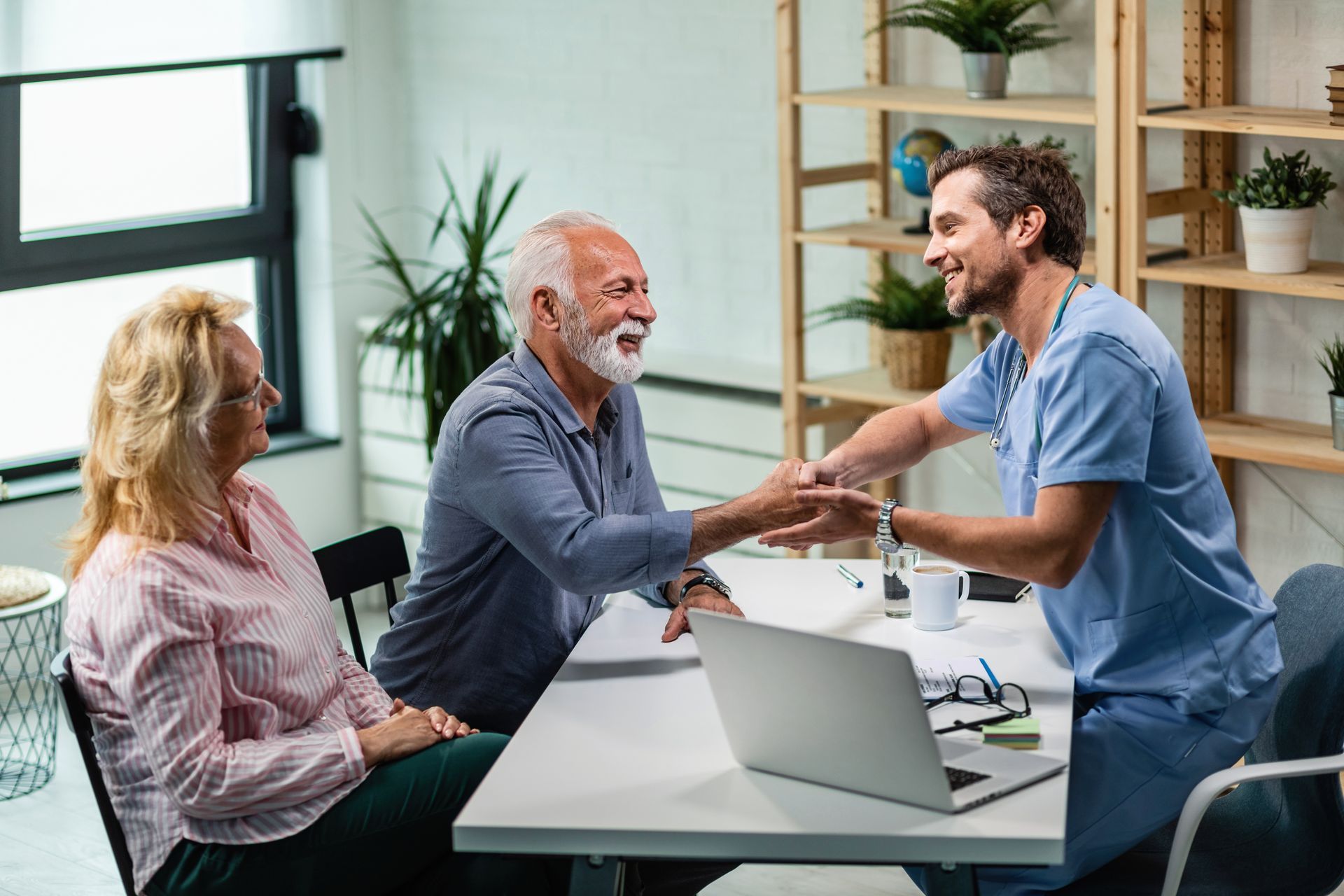 A doctor shakes hands with an elderly man in a medical office. An older woman sits beside the man, watching.