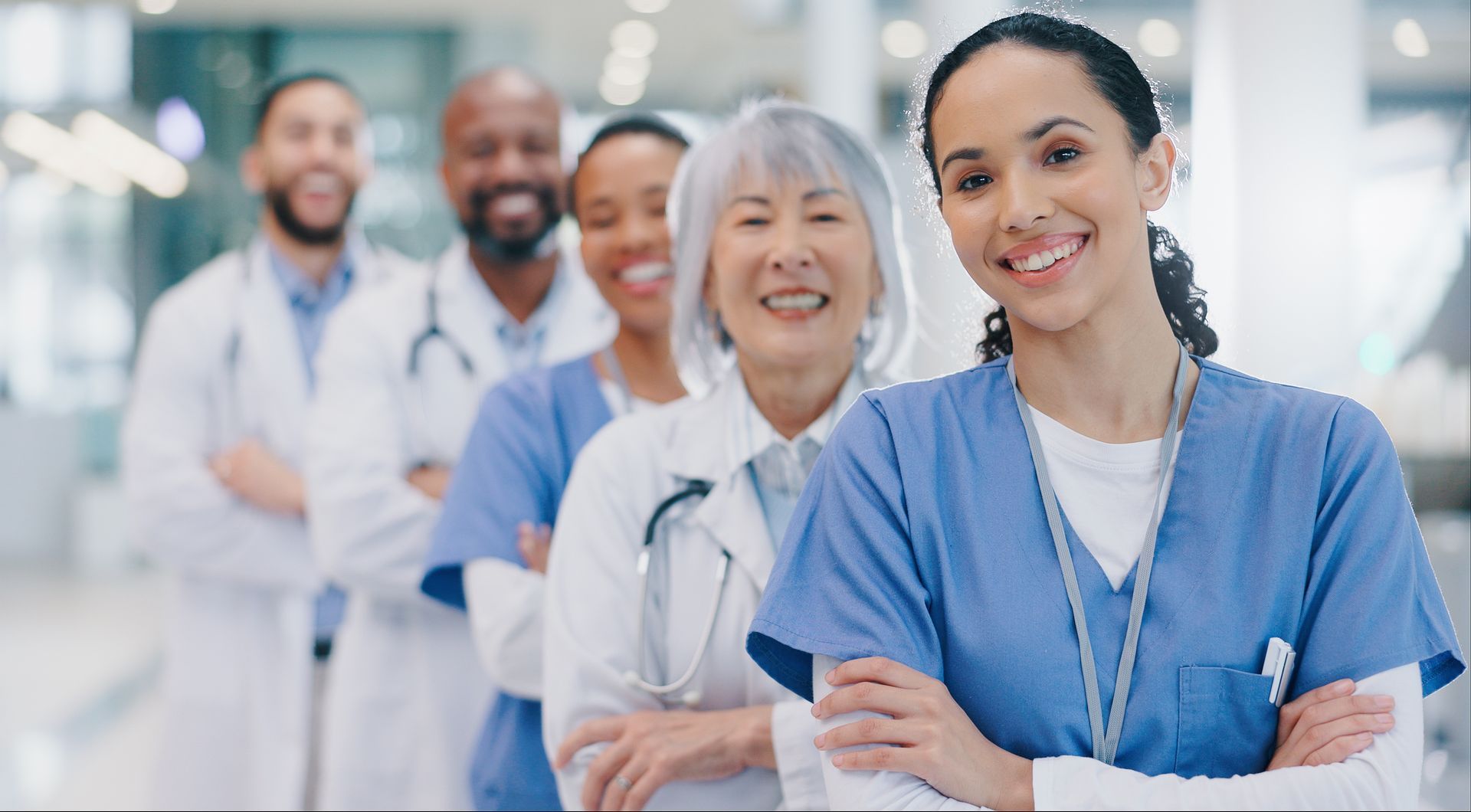 Group of diverse medical professionals, in scrubs and white coats, smiling and standing with arms crossed in a bright hospital hallway.