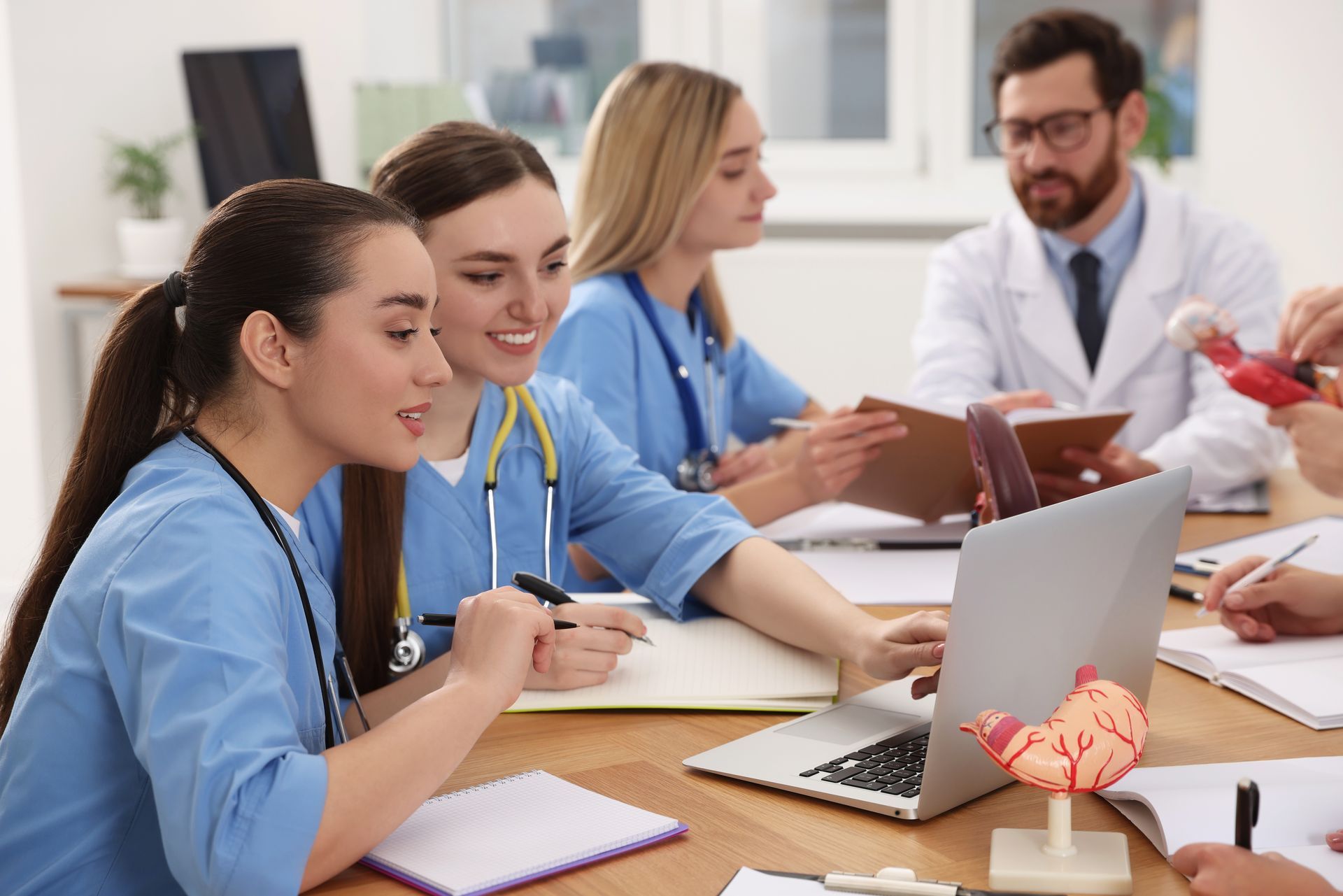 Medical students collaborating around a table, looking at a laptop, with a model organ and notes; a doctor oversees.