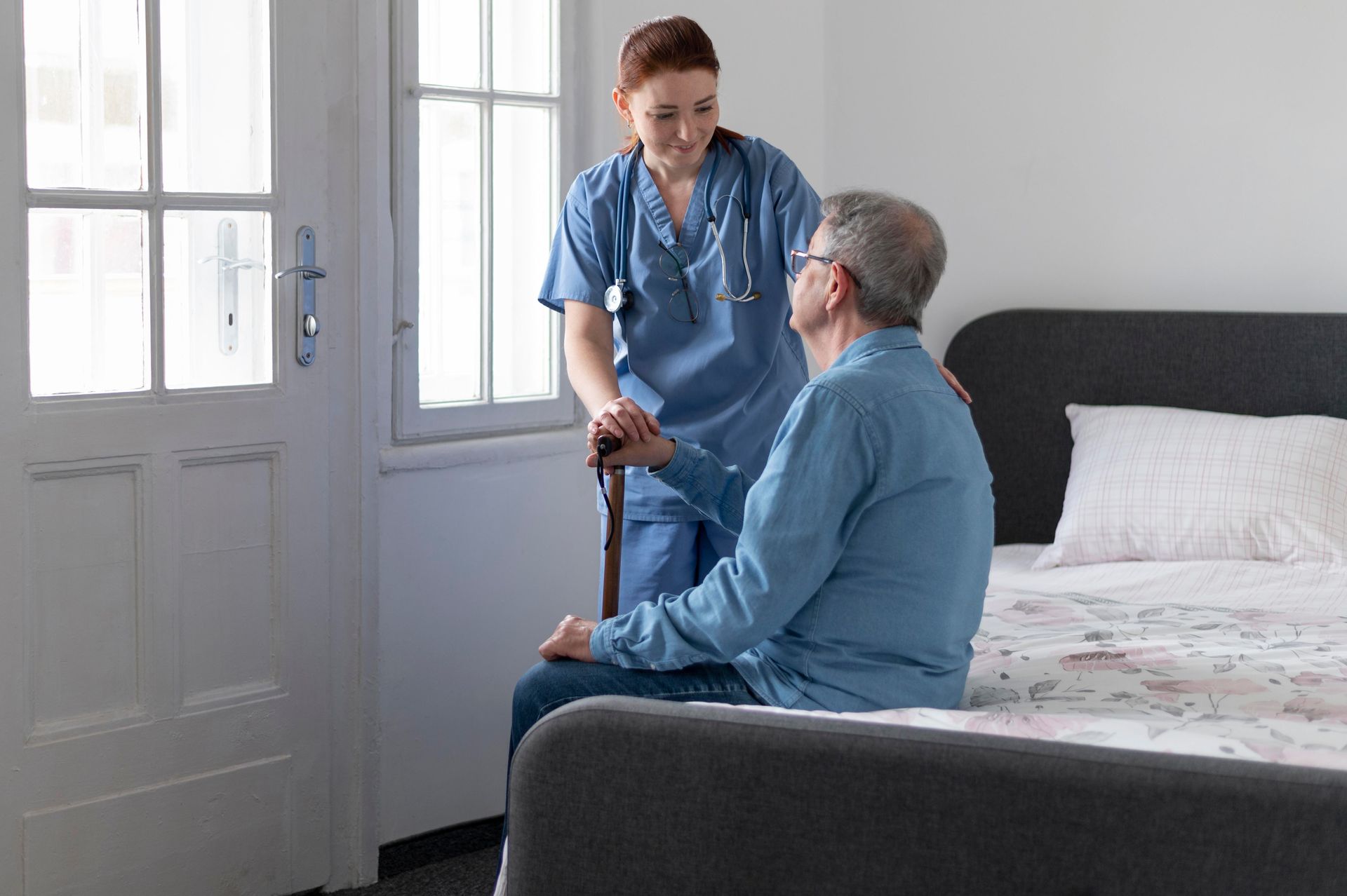 Nurse in blue scrubs assists an elderly man sitting on a bed, holding his cane. She smiles, standing by a window in a room.