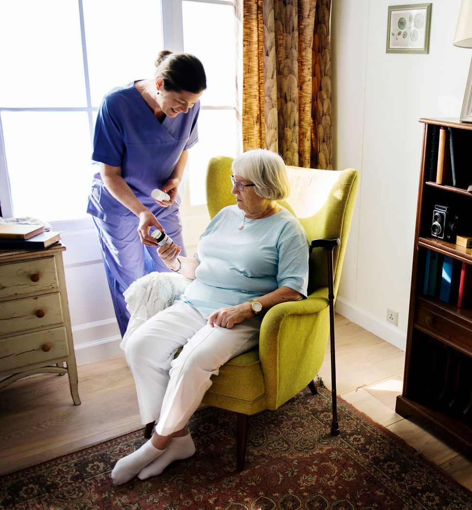 A healthcare worker in blue scrubs assists an elderly woman seated in a yellow armchair. The woman has a cane and is wearing white pants.