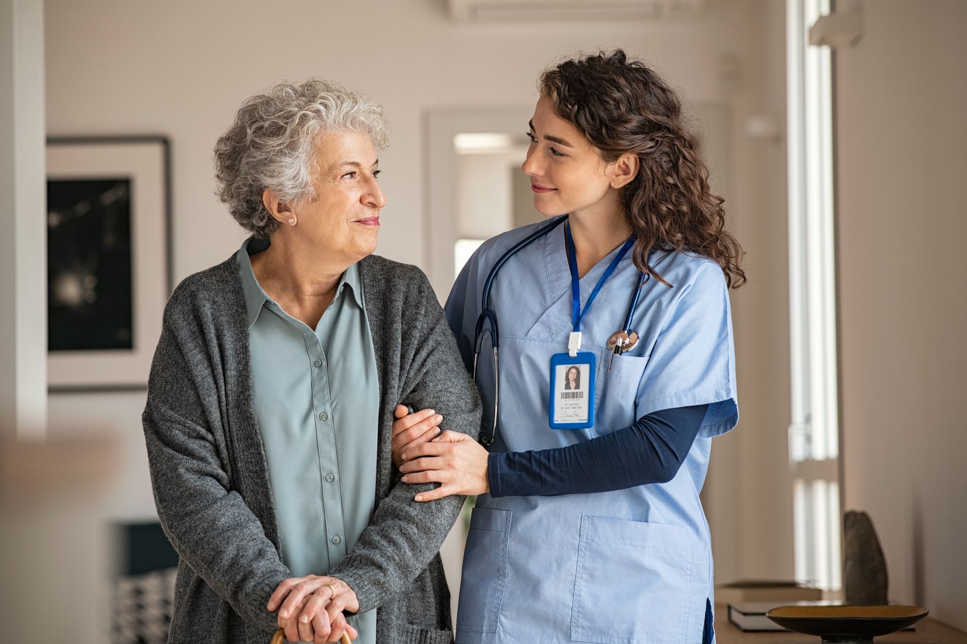 A young female nurse in blue scrubs assists an elderly woman with gray hair as they walk down a hallway.