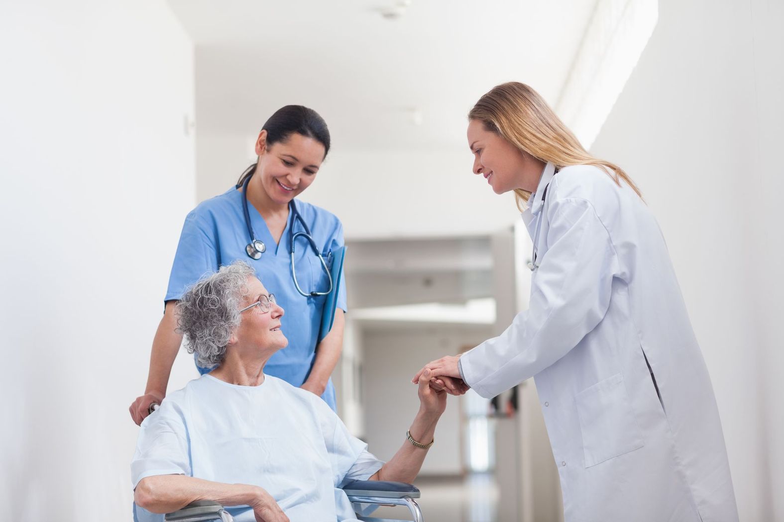 An elderly woman in a wheelchair smiles at a doctor who holds her hand; a nurse stands beside them in a hospital hallway.