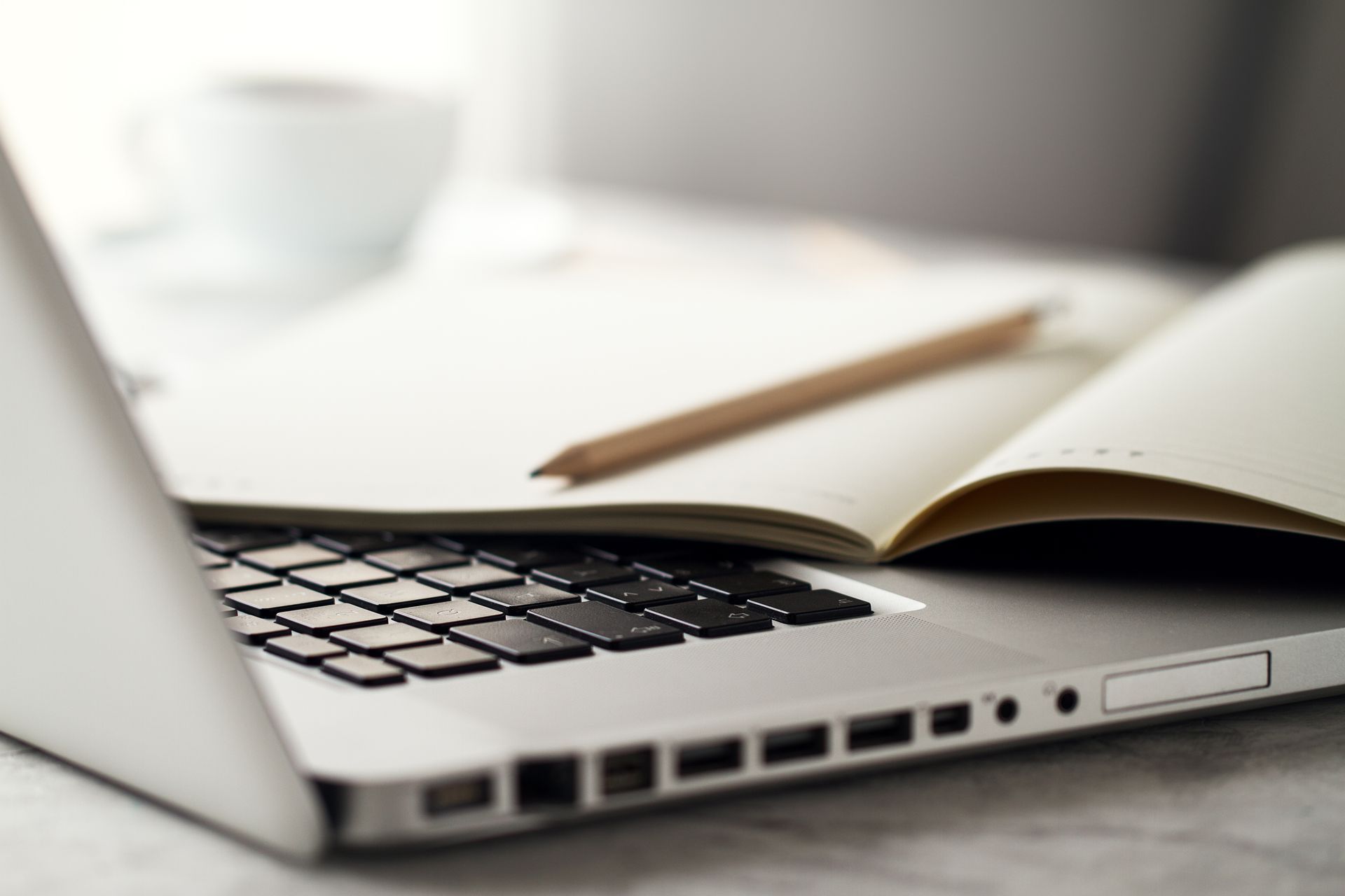 Laptop with open notebook and pencil, coffee cup in background, all on a white surface.